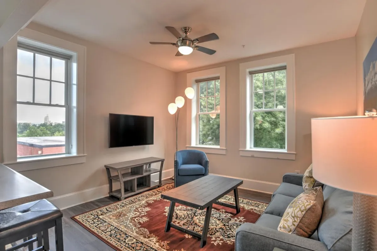 The image showcases a sunlit living area featuring a comfortable sofa, a modern coffee table, a sleek TV, and a stylish floor lamp, all set against large windows that let natural light fill the space. The scene radiates warmth and coziness, inviting relaxation and leisure.