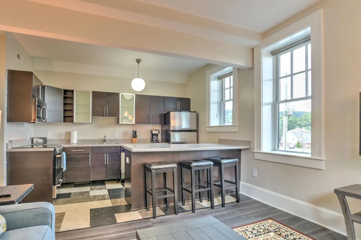 This image showcases a contemporary kitchen with a central island and stools, complemented by stainless steel appliances and windows that bring in natural light. The setting is ideal for both cooking and casual dining, reflecting a space designed for efficiency and shared experiences.