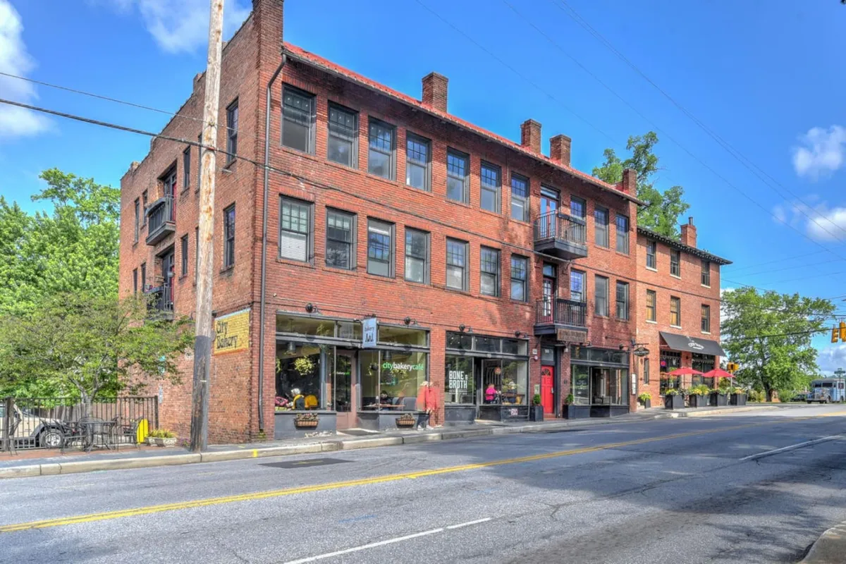 This image captures a lively stretch of historic Charlotte Street, featuring a charming brick facade with large windows and a welcoming balcony above bustling storefronts. Pedestrians can be seen shopping, dining at outdoor cafes, and soaking in the vibrant street atmosphere. The overall scene buzzes with activity and community spirit, highlighting both the architectural character and social energy of the area.