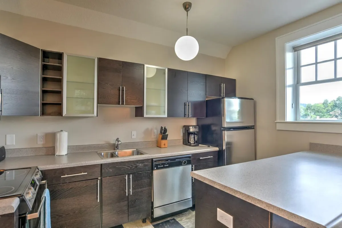 A bright, modern kitchen focused on functional cabinetry, stainless steel appliances, and a kitchen island. A window offers a pleasant view and brings in a sense of calm, with hints of the space being used for baking, brunching, or simply enjoying a peaceful moment.