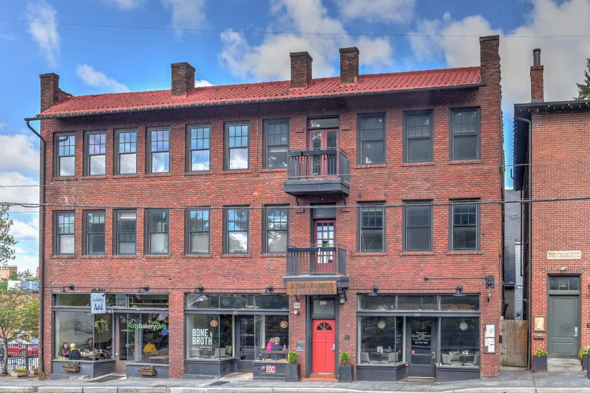 The scene showcases another exterior view along Charlotte Street, focusing on a building with a distinctive red door and large windows. People are seen shopping at local stores, enjoying coffee at nearby cafes, and engaging in casual conversation, reinforcing the sense of neighborhood community. Hints of local art displays and a seasonal market add color and authenticity to the setting.