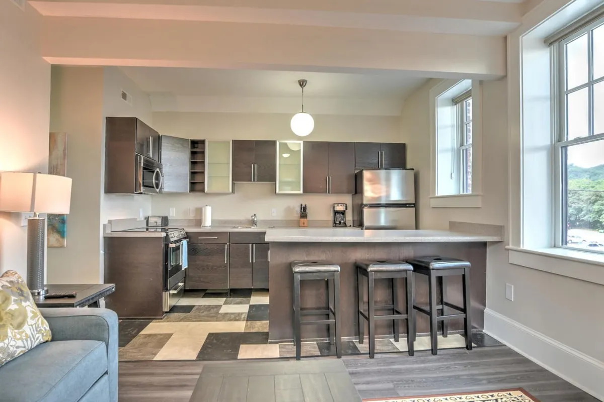 A bright, contemporary kitchen scene with a central island, comfortable stools, and sleek stainless steel appliances. Sunlight pours in through large windows, illuminating the space and creating a welcoming atmosphere for casual dining or entertaining.