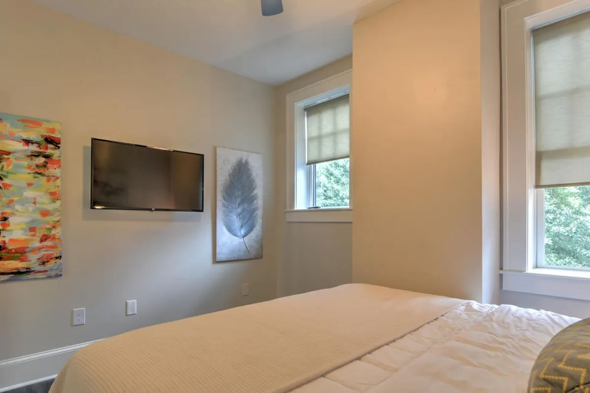 A bright bedroom with a comfortable bed dressed in light bedding, complemented by colorful abstract and leaf artwork. A wall-mounted television provides added convenience, while sunlight brightens the space, creating a welcoming, restful environment.